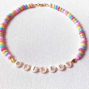May include: A beaded necklace featuring a repeating pattern of small, multi-colored, faceted beads in shades of pink, yellow, blue, and green. The center includes white circular beads spelling the name BRINLEY with small gold-toned heart accents.