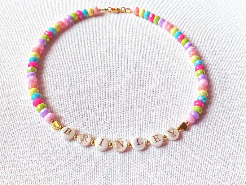 May include: A beaded necklace featuring a repeating pattern of small, multi-colored, faceted beads in shades of pink, yellow, blue, and green. The center includes white circular beads spelling the name BRINLEY with small gold-toned heart accents.