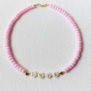 May include: A beaded necklace featuring light pink faceted beads, small gold-toned spacer beads, and four white circular beads with the black letters E, V, I, and E printed on them. The necklace includes two small gold-toned heart charms.