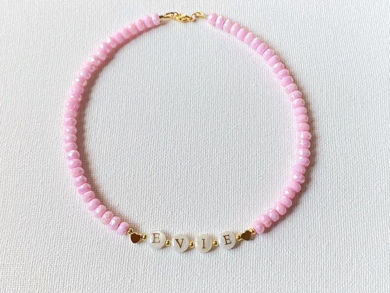 May include: A beaded necklace featuring light pink faceted beads, small gold-toned spacer beads, and four white circular beads with the black letters E, V, I, and E printed on them. The necklace includes two small gold-toned heart charms.