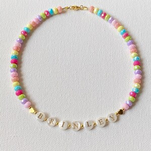 May include: A beaded necklace featuring a repeating pattern of small, multi-colored, faceted beads in shades of pink, yellow, blue, green, and purple. The center displays white circular beads spelling the name BRINLEY with gold-tone accents.