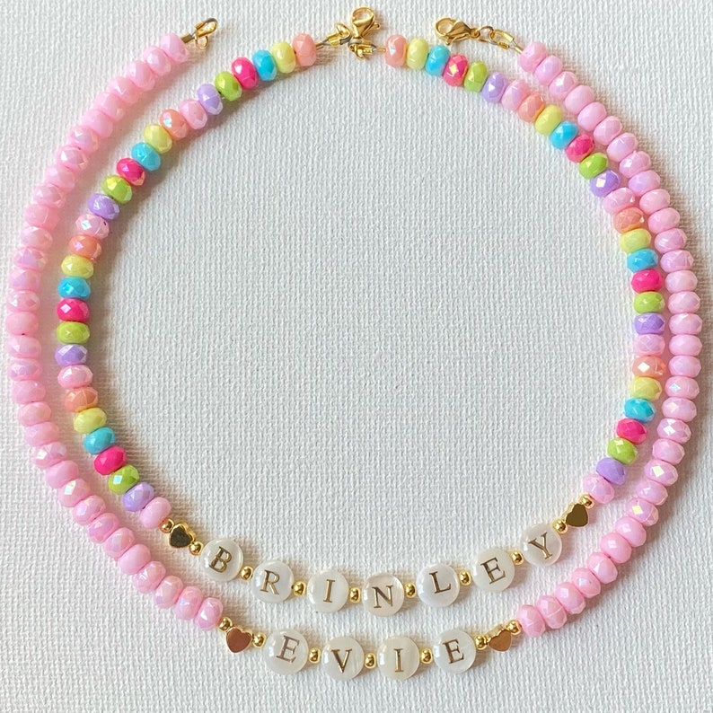 May include: Two beaded necklaces featuring faceted pink beads interspersed with multicolored beads. One necklace displays the name BRINLEY and the other EVIE on white circular beads with gold lettering, accented by small gold heart-shaped charms.