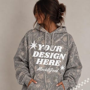May include: A gray and brown camouflage print hooded sweatshirt featuring a front kangaroo pocket and long sleeves. The text "YOUR DESIGN HERE" is printed in large white letters on the chest, accompanied by two white star icons and the text "MockitGood".
