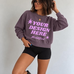 Könnte beinhalten: Ein dunkelviolettes Sweatshirt mit Rundhalsausschnitt und dem hellrosa Schriftzug "YOUR DESIGN HERE" in einer fetten, stilisierten Schriftart, ergänzt durch zwei kleine Sternsymbole und den kursiven Text "MockitGood" auf der Vorderseite.