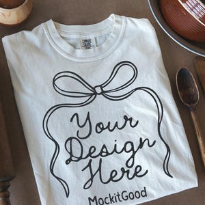 May include: Cream-colored t-shirt with a black bow design and the text "Your Design Here" and "MockitGood". A wooden rolling pin, spoon, and bowl are also in the image. The shirt is laid flat on a brown surface.