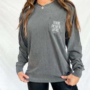 Puede incluir: Una camiseta de manga larga de color gris carbón con un bolsillo en el pecho izquierdo. El bolsillo tiene el texto "YOUR DESIGN HERE" impreso en blanco.