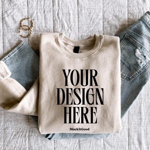 Pode incluir: Uma camisola bege com o texto "YOUR DESIGN HERE" em preto, combinada com jeans azul claro e brincos de argola dourados. A camisola está dobrada e os jeans são desgastados.