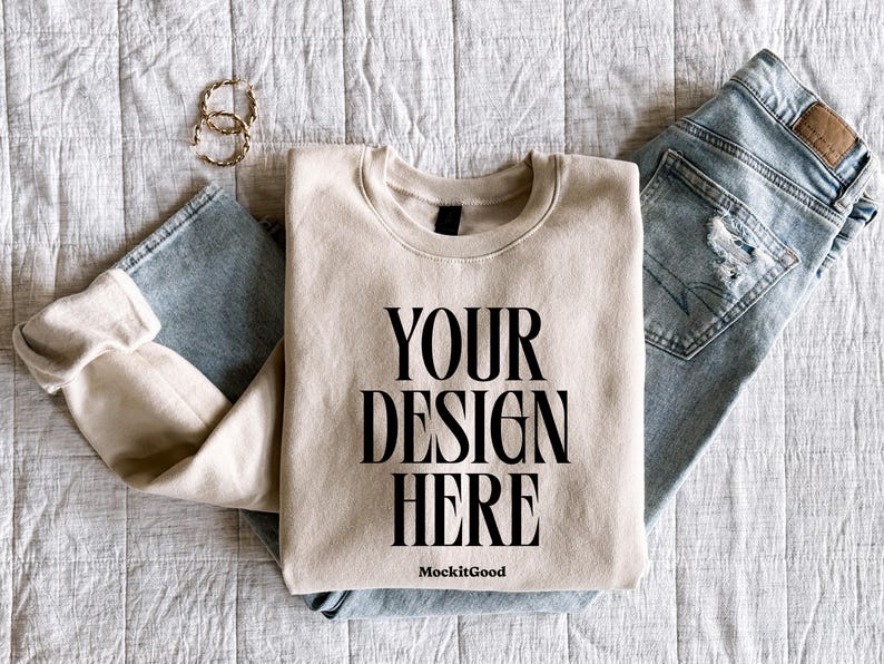 May include: A beige sweatshirt with the text "YOUR DESIGN HERE" in black, paired with light blue jeans and gold hoop earrings. The sweatshirt is folded, and the jeans are distressed.