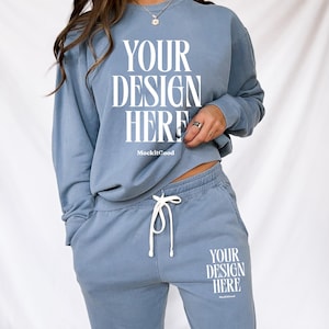 以下が含まれることがあります： ライトブルーのスウェットシャツと、お揃いのスウェットパンツのセット。スウェットシャツには、白いブロック体で「YOUR DESIGN HERE」の文字がプリントされています。スウェットパンツにも同じ文字がプリントされています。セットはドローストリングウエストで、柔らかい素材で作られています。