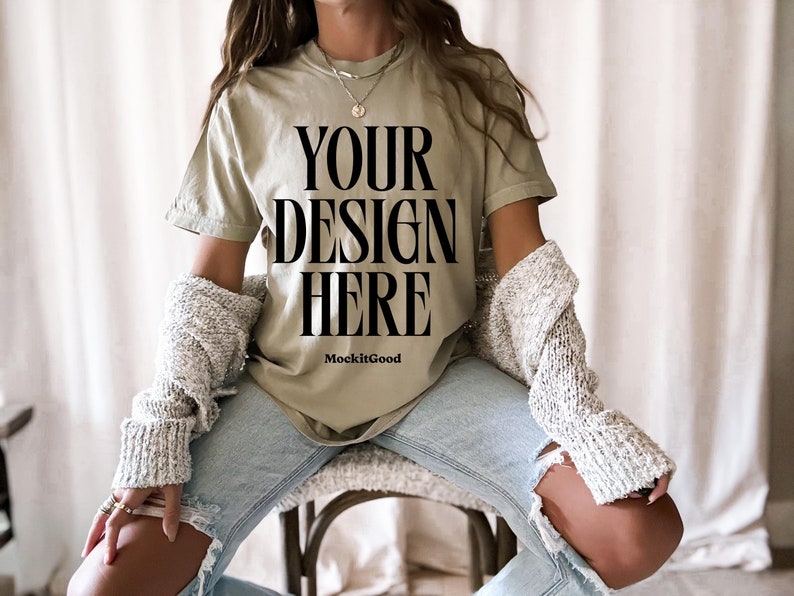 Puede incluir: Una mujer lleva una camiseta verde claro con el texto "YOUR DESIGN HERE" y las palabras "MockitGood" debajo. Lleva pantalones vaqueros azules y una chaqueta blanca de punto.