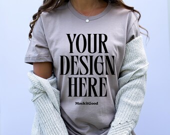May include: A taupe t-shirt featuring the text "YOUR DESIGN HERE" in large, bold, black letters. The text "MockitGood" is printed below the main text. A light grey knit sweater is draped over the shoulders.