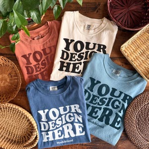 以下が含まれることがあります： テラコッタ、クリーム、ブルーの4枚のTシャツ。それぞれに太字で「YOUR DESIGN HERE」と書かれています。木製の表面には、編まれたバスケットと鉢植えの植物がシャツを囲んでいます。シャツはMockitGood製です。