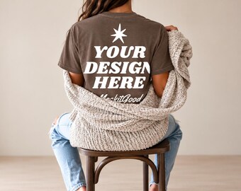 May include: A brown short-sleeve t-shirt featuring a white star icon above the bold white text "YOUR DESIGN HERE" and the script text "MockitGood" printed on the back, worn with a beige knit cardigan and light blue denim jeans.