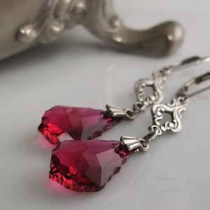May include: A pair of silver dangle earrings with a teardrop-shaped, deep pink crystal on each earring. The earrings have a delicate, ornate design.