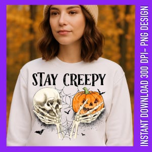 Op de afbeelding: Wit sweatshirt met de tekst "STAY CREEPY" boven een schedel en een pompoen die door skeletachtige handen wordt vastgehouden. Het ontwerp bevat spinnenwebben en vleermuizen, perfect voor Halloween.