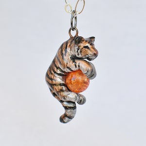 May include: A pendant necklace featuring a tiger figurine holding an orange sphere. The tiger has brown and black stripes with silver accents. The necklace chain is silver.