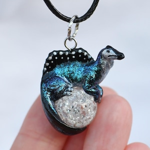 May include: A blue and black dinosaur pendant necklace with a white crystal sphere. The dinosaur is curled around the sphere and has a black and white spiky back.
