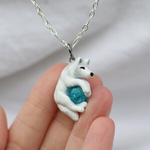 Op de afbeelding: Een ketting met een witte wolf hanger die een blauwe edelsteen in zijn poten vasthoudt.