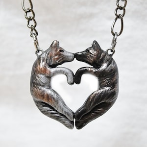 May include: A silver necklace with a pendant of two wolves forming a heart shape. The wolves are facing each other and their noses are touching. The pendant is made of metal and has a textured finish.