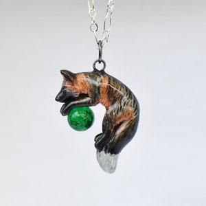 Puede incluir: Un collar de cadena de plata con un colgante de un zorro negro, marrón y blanco que sostiene una bola de cristal verde.