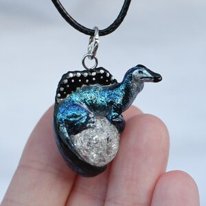 May include: A blue and black dinosaur pendant necklace with a glittery finish. The dinosaur is sitting on a white egg with a glittery finish.