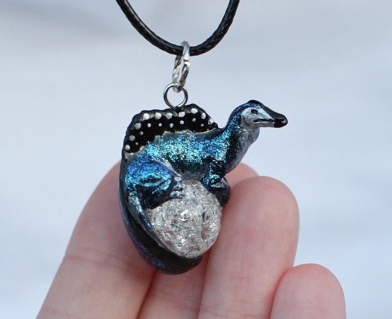 May include: A blue and black dinosaur pendant necklace with a glittery finish. The dinosaur is sitting on a white egg with a glittery finish.