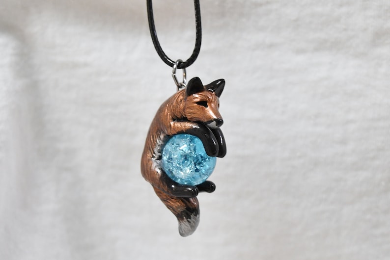 May include: A pendant necklace featuring a fox figurine holding a blue crystal ball. The fox is brown with black accents and a silver tail. The crystal ball is a light blue color and appears to be made of glass. The necklace has a black cord.