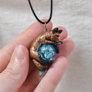 May include: A handcrafted pendant necklace featuring a fox figurine embracing a blue crystal ball. The fox is detailed with brown and black accents, and the crystal has a cracked appearance. The pendant hangs from a black cord.