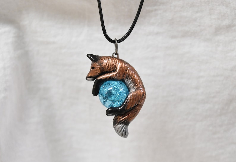 May include: A pendant necklace featuring a brown fox figurine embracing a blue crystal ball. The fox has black accents and a silver-colored bail. The necklace has a black cord.