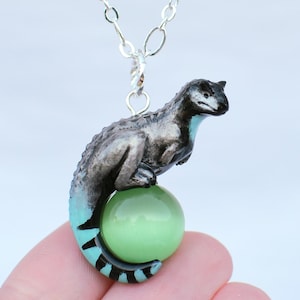 May include: A silver necklace with a dinosaur pendant. The dinosaur is grey and black, with a turquoise tail, sitting on a light green orb. The necklace chain is silver.
