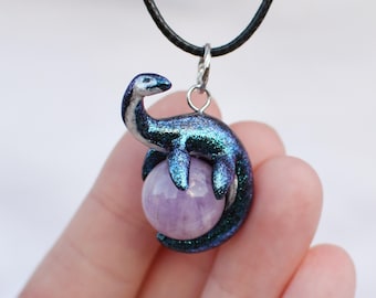 May include: A silver necklace with a black cord and a pendant featuring a blue and green iridescent dinosaur figurine perched on a round, light purple gemstone.