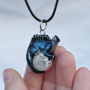 May include: A black cord necklace with a blue and black iridescent polymer clay pendant featuring a small lizard curled around a white crystal ball.