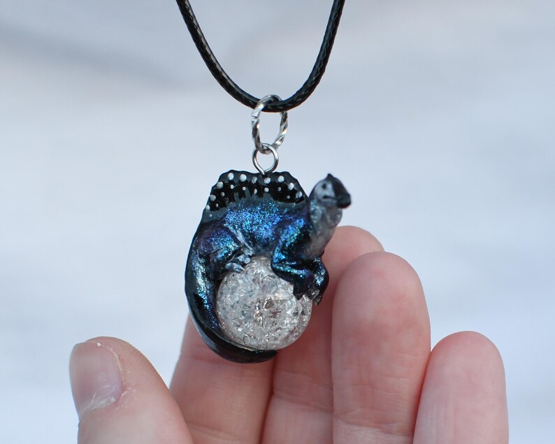 May include: A black cord necklace with a blue and black iridescent polymer clay pendant featuring a small lizard curled around a white crystal ball.