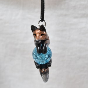 May include: A pendant featuring a fox figurine with copper-colored fur and black accents, holding a blue, cracked-glass orb. The fox is suspended from a black cord with a silver-colored clasp, creating a whimsical and unique accessory.