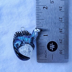 May include: A blue and black dinosaur charm with white spots, sitting on a clear crystal egg. The charm is hanging from a silver loop.