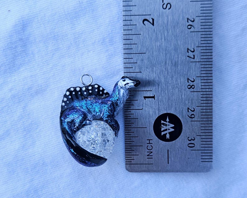 May include: A blue and black dinosaur charm with white spots, sitting on a clear crystal egg. The charm is hanging from a silver loop.