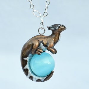 May include: A silver chain necklace with a bronze dragon pendant holding a light blue gemstone. The dragon is curled around the gemstone, with its tail wrapped around the bottom.