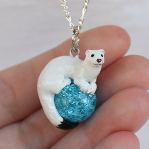 May include: A white ceramic necklace pendant featuring a small white weasel or stoat figurine perched on a blue sparkly ball.