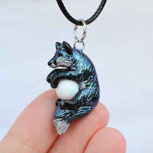 Puede incluir: Un collar con un colgante de zorro azul y negro con una piedra de ópalo blanca. El zorro está enroscado y sostiene el ópalo en sus patas.
