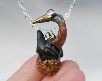 May include: A handcrafted pendant necklace with a bird design. The bird has a brown body, black accents, and a gold beak. The pendant is attached to a silver chain. The bird is perched on a textured, amber-coloured sphere.