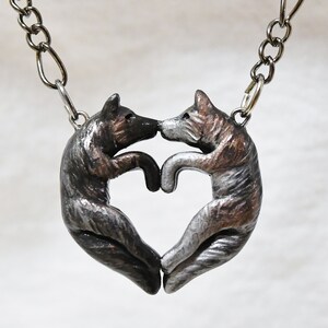 May include: A silver necklace with a heart-shaped pendant featuring two wolves facing each other. The wolves are sculpted in a realistic style and have a silver and bronze finish.