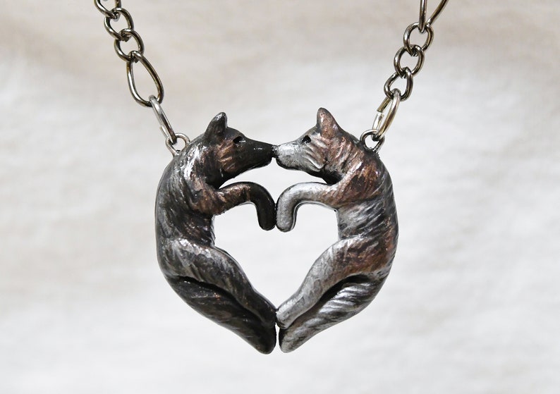 May include: A silver necklace with a heart-shaped pendant featuring two wolves facing each other. The wolves are sculpted in a realistic style and have a silver and bronze finish.