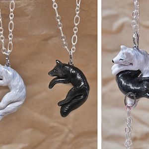 May include: Two silver necklaces with wolf pendants. One pendant is a white wolf curled up, the other is a black wolf curled up. The wolves are facing each other.