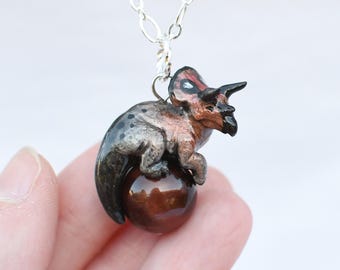 May include: A pendant necklace with a miniature triceratops dinosaur in brown, black, and red. The dinosaur sits on a brown, spherical bead. The pendant hangs from a delicate silver chain, creating a unique accessory. The necklace is a charming piece of jewellery.