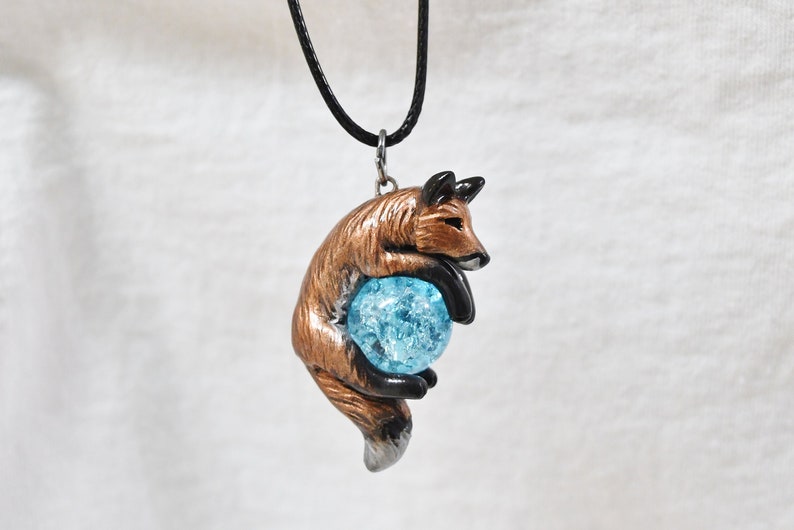 May include: A pendant necklace featuring a brown and black fox figurine holding a blue, cracked-glass orb. The fox is suspended from a black cord. The pendant is a unique piece of jewelry.