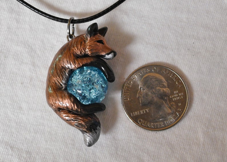 May include: A handcrafted fox pendant, holding a blue crystal ball. The fox is primarily brown with black and silver accents. The pendant hangs from a black cord. A U.S. quarter is next to the pendant for size reference.