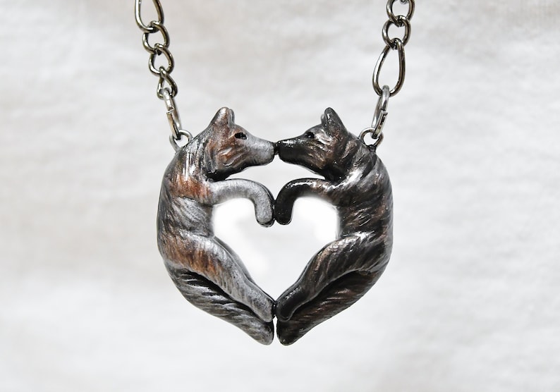 May include: A silver necklace with a pendant of two wolves forming a heart shape. The wolves are facing each other and their noses are touching. The pendant is made of metal and has a textured finish.