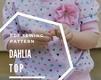 May include: A white and pink children's top with a colorful heart print. The top has a pink flower embellishment and the text "PDF SEWING PATTERN DAHLIA TOP GOLDENRIPPY.COM/SHOP" is visible.