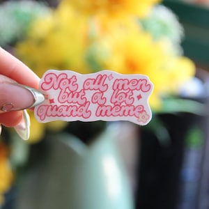 Peut inclure: Un autocollant rose et blanc avec le texte "Not all men but a lot quand même" est tenu devant un arrière-plan flou de fleurs jaunes et d'un vase vert. L'autocollant a un motif floral.