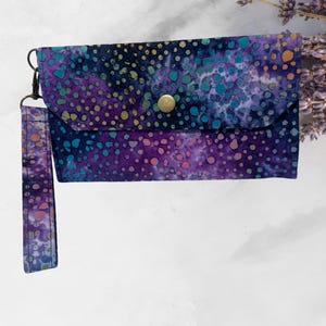 May include: A purple and blue fabric wallet with a gold button closure and a matching strap. The wallet has a tie-dye pattern with white and yellow dots.